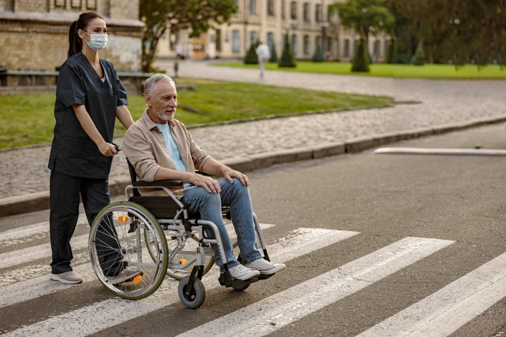 Full length shot of young nurse in protective face mask taking care of mature disabled man in